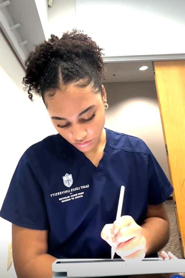 Jordan Ireland, a female student with curly hair wearing a pair of Saint Louis University scrubs, writes on a tablet